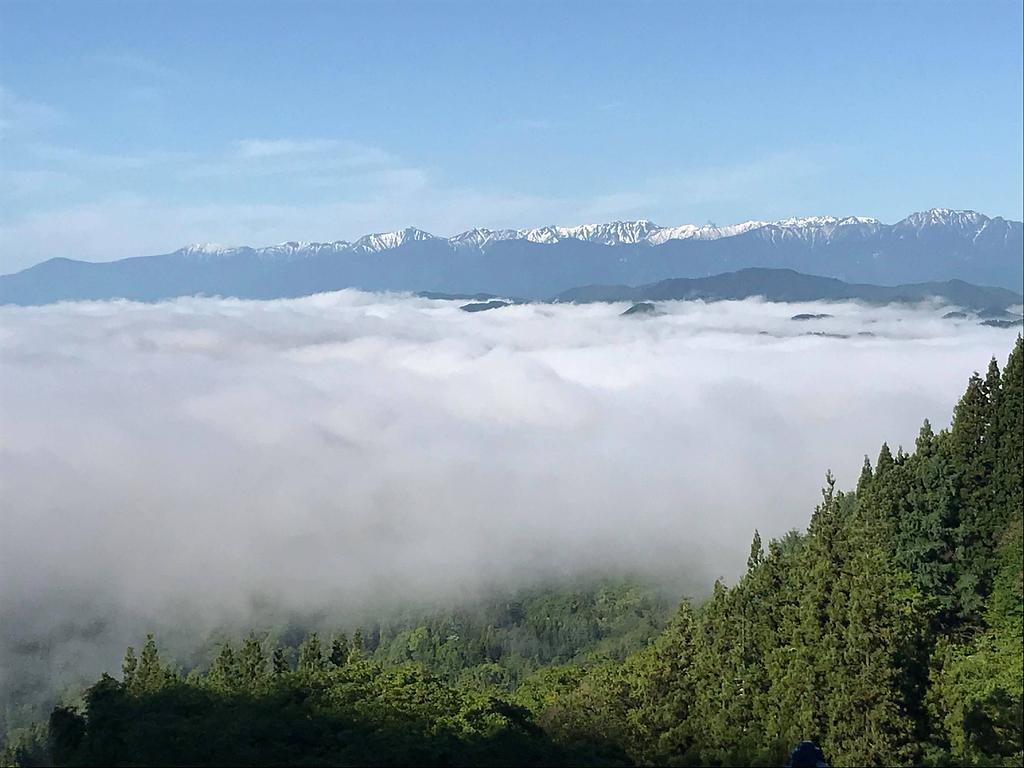 山頂から見下ろす広大な雲海と、遠くにそびえる雪をかぶった山々の景色。手前には緑豊かな森林が広がり、自然の壮大さと静けさを感じさせる光景。