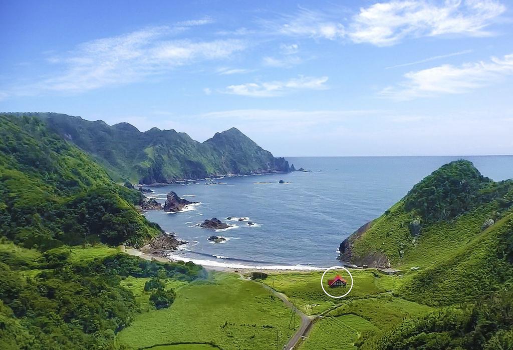 緑豊かな山々に囲まれた海岸の風景。遠くに広がる青い海と山々の間に、赤い屋根の家が小さく見える。自然の中にひっそりと佇む家が強調されており、静寂で美しい環境が広がる。