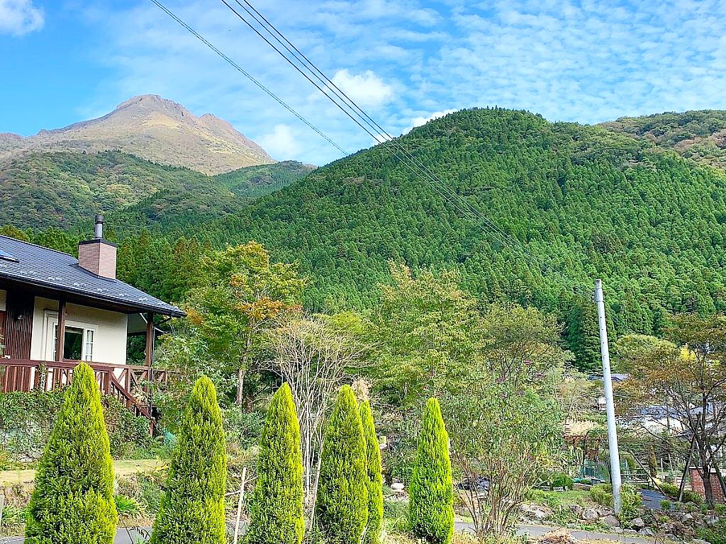 青空の下、緑豊かな山々に囲まれた家の風景。手前には整えられた庭木が並び、奥には深い緑の山が広がる。左側には煙突のある家があり、自然に囲まれた静かな環境が広がっている。