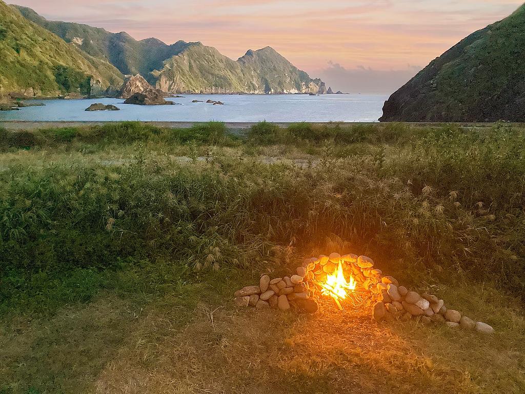 夕暮れ時、海を望む草原に設置された小さな焚き火。周囲には石で囲まれた焚き火台があり、穏やかな炎が揺らめいている。背景には壮大な海と山々が広がり、自然の静けさと美しさを感じられる風景。