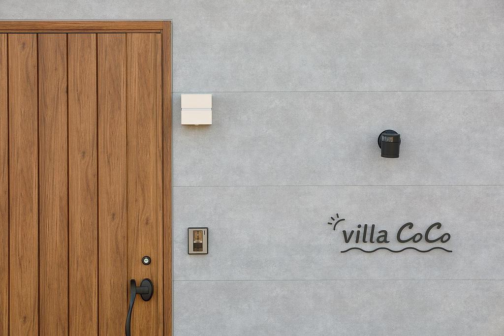 モダンなデザインの玄関。木製のドアとグレーの壁に、スタイリッシュな「villa CoCo」のサインが施されている。シンプルながらも洗練された外観が特徴的。