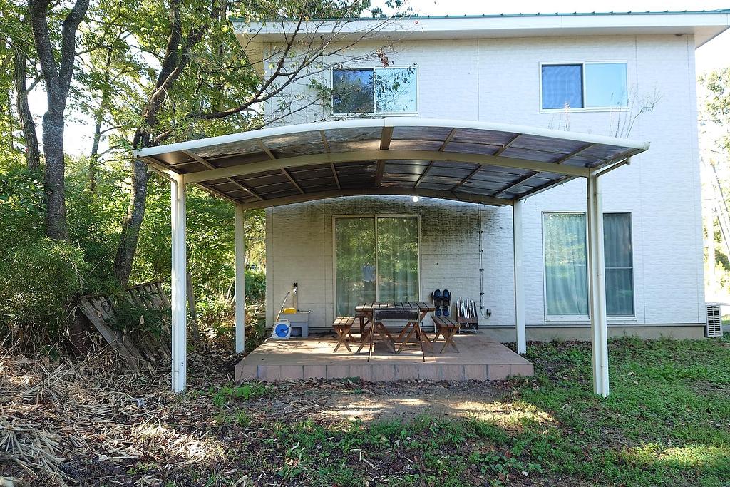 緑豊かな庭に面した白い2階建ての建物の裏側にある屋根付きのバーベキューテラス。木製のテーブルと椅子が設置されており、自然の中でバーベキューを楽しむことができる。