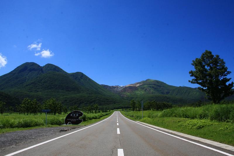 青空の下、まっすぐに続く道路の先に緑豊かな山々が広がる風景。道路の両側には草原が広がり、遠くに山頂が見える。自然の中を走る開放感あふれる景色。