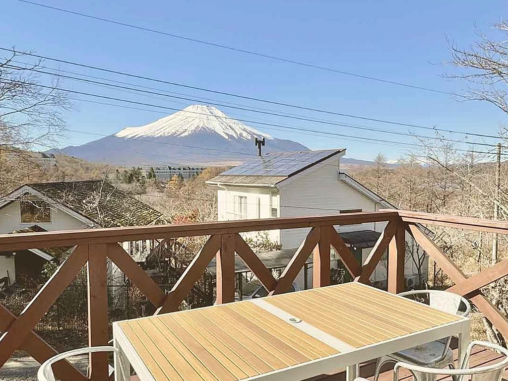 木製のテラスから富士山を眺める景色。手前にはアウトドア用のテーブルと椅子が設置され、背景には雪を冠した富士山が青空の下に雄大にそびえている。