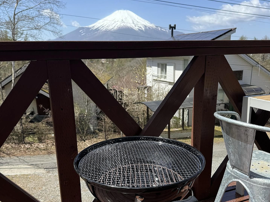 ウッドデッキに設置されたバーベキューグリル越しに、遠くに富士山が見える風景。晴れた空と山の白い雪、周囲の木々や建物が調和した景色が広がっている。